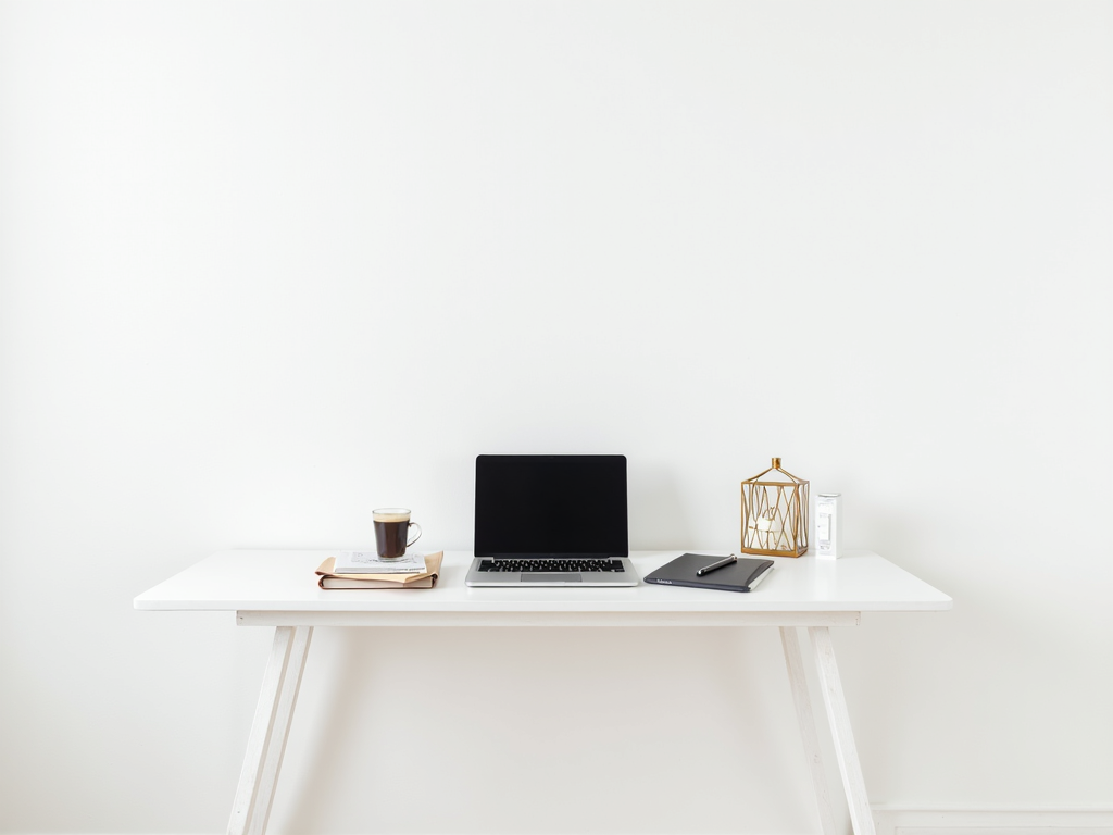 Ein sauberer Schreibtisch mit nur einem Laptop, einer Kaffeetasse und einem Notizbuch vor einer schlichten weißen Wand.