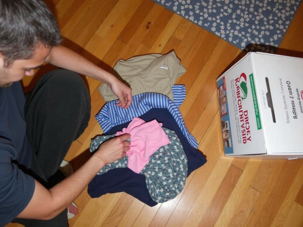 Eine Person sortiert einen kleinen Kleiderstapel auf einem Holzboden, mit einer Spendenbox in der Nähe.