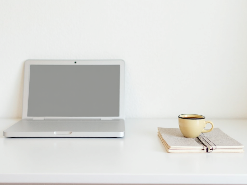 Ein sauberer, einfacher Schreibtisch mit nur einem Laptop, einer Tasse Kaffee und einem Notizblock vor einer weißen Wand.